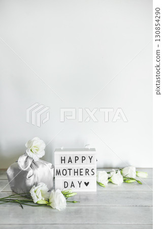 Light box with lettering Happy Mother's Day, Gift box trendy wrapped in silk fabric in Furoshiki technique, white flowers Eustoma or Lisianthus on light wooden background with copy space. Light box with lettering Happy Mother's Day, Gift box trendy wrapped in silk fabric in Furoshiki technique, white flowers Eustoma or Lisianthus on light wooden background with copy space. 130854290