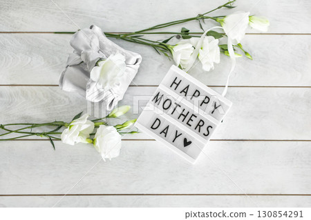 Light box with lettering Happy Mother's Day, Gift box trendy wrapped in silk fabric in Furoshiki technique, white flowers Eustoma or Lisianthus on light wooden background with copy space. Light box with lettering Happy Mother's Day, Gift box trendy wrapped in silk fabric in Furoshiki technique, white flowers Eustoma or Lisianthus on light wooden background with copy space. 130854291