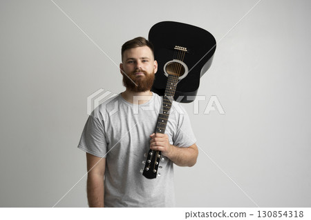 Bearded young man in grey t-shirt holding black acoustic guitar on shoulder in studio 130854318