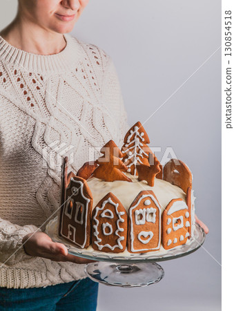 Woman holds homemade cake with gingerbread city house, Christmas tree and reindeer on glass stand 130854518