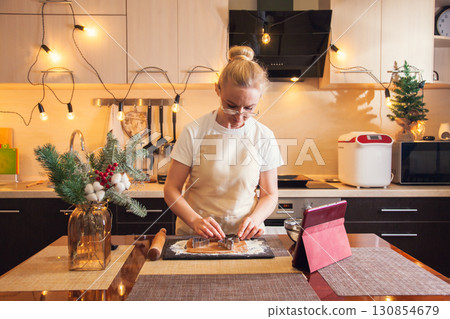 Woman following recipe on digital tablet and cuts out dough with mold for cooking christmas gingerbread cookies Woman following recipe on digital tablet and cuts out dough with mold for cooking christmas gingerbread cookies 130854679