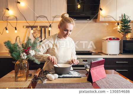 Woman in christmas sweater following recipe on digital tablet and cooking dough on her kitchen Woman in christmas sweater following recipe on digital tablet and cooking dough on her kitchen 130854686