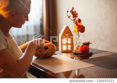 Woman carves pumpkin for Halloween in room with autumn decor and lamp house. Cosy home and preparing for Halloween. Woman carves pumpkin for Halloween in room with autumn decor and lamp house. Cosy home and preparing for Halloween. 130855521