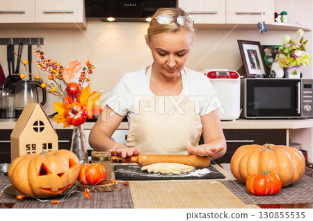 A woman rolls out cookie dough for Halloween with a rolling pin in the kitchen with an autumn decor. A woman rolls out cookie dough for Halloween with a rolling pin in the kitchen with an autumn decor. 130855535