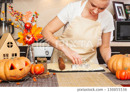 A woman cuts out cookies for Halloween with a mold in the kitchen with an autumn decor. 130855536