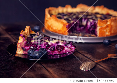 Rustic shortbread pie with blueberries in sour cream filling on wooden background with piece cut off on saucer and spoon 130855769