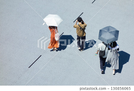 Midsummer afternoon sun and tourists carrying parasols walking across a city square 130856065