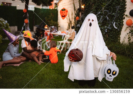 Child wearing ghost costume holding pumpkin and skull trick or treat bag during Halloween party in the garden 130856159