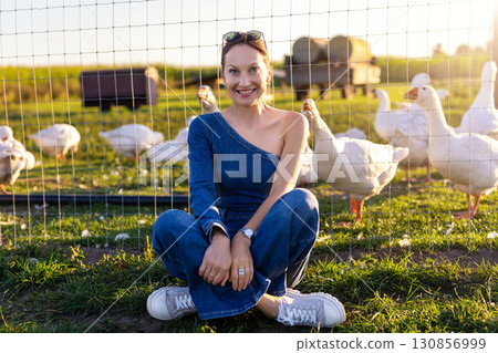 Portrait young adult happy smiling woman wear jeans denim jumpsuit enjoy having fun countryside farm animal geese flock warm sunset light. Female person rural country village farmyard bird poultry 130856999