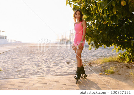 Young beauty woman in pink sportswear on rollerblades near the beach at golden sunset 130857151