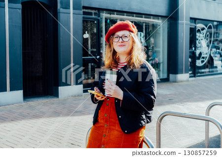 Young smiling plus size woman with backpack using mobile phone, drinking coffee outdoors on the street. Confident fashion hipster messaging, chatting, working remotely. Using navigator app for travel. 130857259