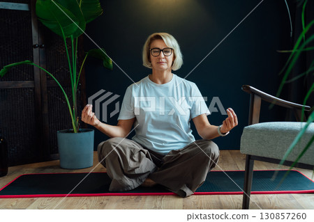 Middle aged woman practicing meditation at home, sitting in lotus position on floor yoga mat in modern Scandi dark interior home. Meditating, relaxed, eyes closed. Mental health and well being. Middle aged woman practicing meditation at home, sitting in lotus position on floor yoga mat in modern Scandi dark interior home. Meditating, relaxed, eyes closed. Mental health and well being. 130857260