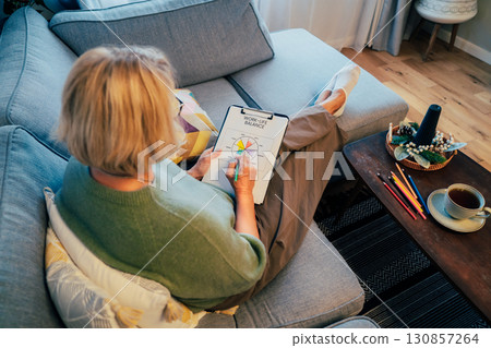 Woman drawing work-life balance wheel sitting on the sofa at home. Self-reflection and life planning for next new year. Personal Year review. Coaching tools. Finding Balance in Life. Selective focus. 130857264