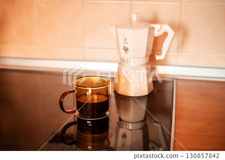 Kyiv, Ukraine. 24 April 2025 A brown glass mug filled with coffee sits near a traditional white moka pot on a shiny black surface, ready to be served. Kyiv, Ukraine. 24 April 2025 A brown glass mug filled with coffee sits near a traditional white moka pot on a shiny black surface, ready to be served. 130857842