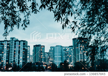 A serene cityscape at dusk, where the illuminated windows of buildings reflect beautifully on the water. Overhanging leaves frame the sky above. A serene cityscape at dusk, where the illuminated windows of buildings reflect beautifully on the water. Overhanging leaves frame the sky above. 130857843