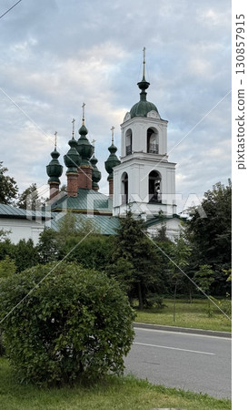 Russian orthodox church with green domes and white bell tower surrounded by trees and bushes 130857915