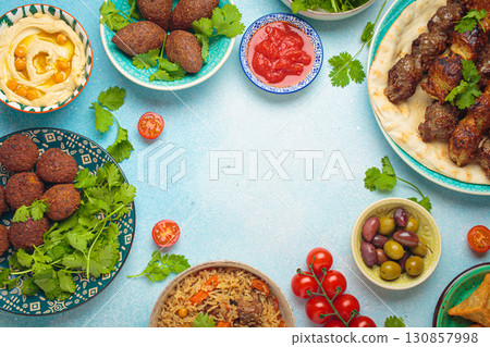 Top view of a vibrant table filled with lebanese dishes and mezze, featuring meat kebabs, falafel, kibbeh, hummus, sambousek, rice, olives. Middle eastern or Arab dishes, space for text Top view of a vibrant table filled with lebanese dishes and mezze, featuring meat kebabs, falafel, kibbeh, hummus, sambousek, rice, olives. Middle eastern or Arab dishes, space for text 130857998