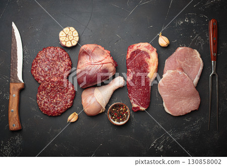 Selection of assorted raw meat cuts for zero carb carnivore diet: uncooked beef steak, pork meat, ground meat patty, heart, and chicken legs on black stone background top view 130858002
