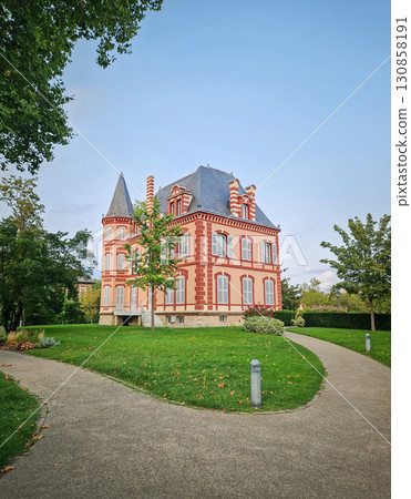 The Castel Rose in L'Isle-Adam, France. Historic mansion characterized by its pink walls and distinctive red brick trim. The building is surrounded by a lush green lawn and winding paths 130858191