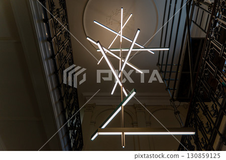 A close-up of a contemporary light fixture featuring multiple intersecting light tubes suspended in a stairwell. A close-up of a contemporary light fixture featuring multiple intersecting light tubes suspended in a stairwell. 130859125