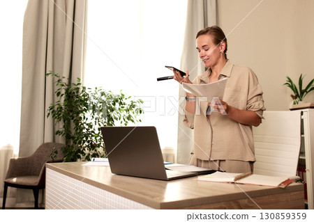Modern professional speaking into voice recorder while reviewing documents at her desk Modern professional speaking into voice recorder while reviewing documents at her desk 130859359