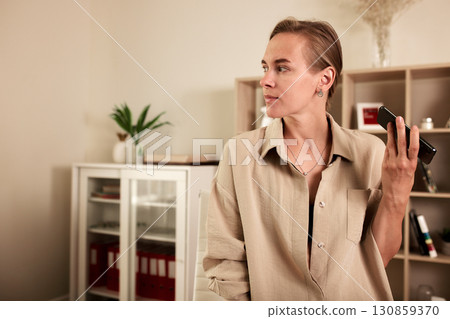 Professional woman holding smartphone while working in modern home office setting 130859370
