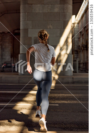 Woman jogging outdoors under an urban bridge in morning sunlight 130859416