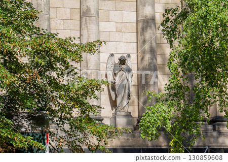 A sculpture of an angel with large wings, carved from stone, is located on the facade of an ancient building between two columns. The figure seems to embrace itself. It is partially hidden by green tr 130859608