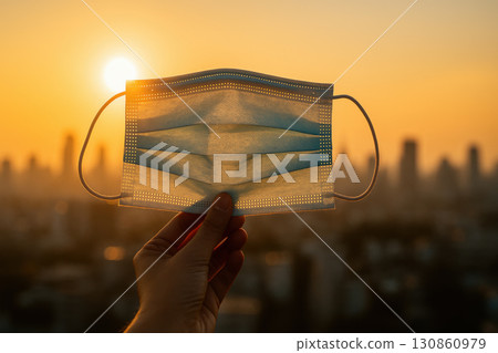 Surgical mask held by hand against bright sunset with city skyline background, symbolizing health care and pandemic safety awareness in urban environment 130860979