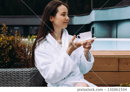 A relaxed woman enjoys cup of tea near a pool, wearing a white robe in a peaceful setting. A serene girl enjoys a moment with a cup of morning coffee. 130861504