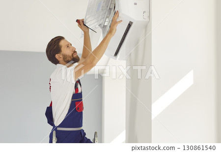 Professional electrician man with screwdriver maintaining modern air conditioner indoors. Professional electrician man with screwdriver maintaining modern air conditioner indoors. 130861540