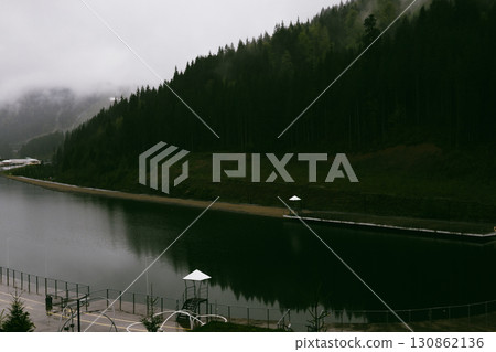 A serene mountain lake reflects the surrounding landscape, with pine trees and structures under a cloudy sky, creating a peaceful atmosphere in autumn 130862136