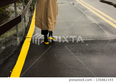 A person in a tan coat walks along a wet, yellow-lined road on a rainy day seen from behind. A woman wearing black boots with yellow soles on a street A person in a tan coat walks along a wet, yellow-lined road on a rainy day seen from behind. A woman wearing black boots with yellow soles on a street 130862143