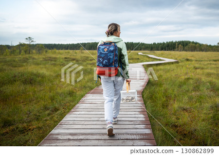 Rearview woman forager moving along marked eco trail in wetland woodland carrying harvest basket 130862899