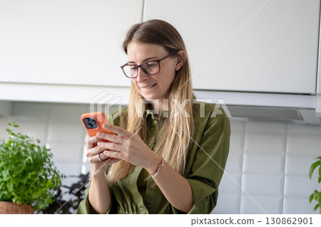 Happy woman texting on smartphone in kitchen in relaxed mood. Social media chat or online shopping 130862901