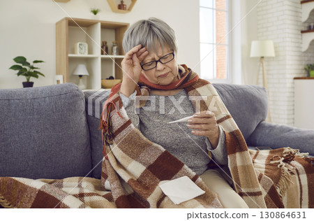 Sick senior woman checking temperature wrapped in a blanket suffering from headache. Sick senior woman checking temperature wrapped in a blanket suffering from headache. 130864631