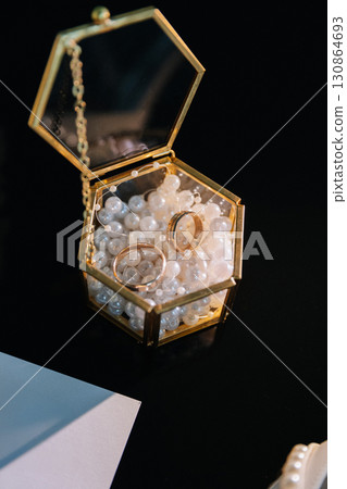 Two gold wedding rings in a glass box filled with decorative pearls on black background 130864693
