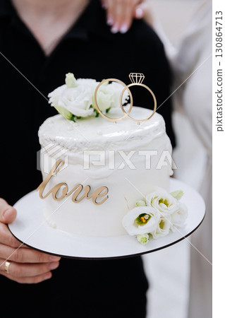 Elegant wedding cake with rings and white flowers, held by newlyweds in celebration Elegant wedding cake with rings and white flowers, held by newlyweds in celebration 130864713