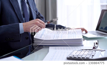Businessman reviewing ring folder of financial documents with magnifying glass at desk in modern office. Audit and taxes in business Businessman reviewing ring folder of financial documents with magnifying glass at desk in modern office. Audit and taxes in business 130864775