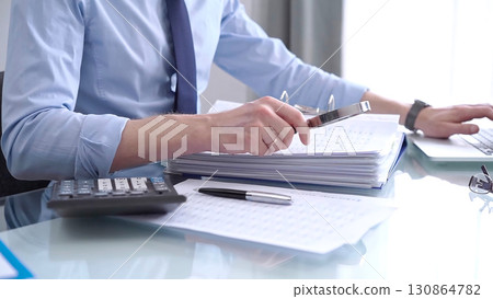 Businessman in blue short is using calculator and reviewing ring folder of financial documents with magnifying glass at desk in modern office. Audit and taxes in business concept Businessman in blue short is using calculator and reviewing ring folder of financial documents with magnifying glass at desk in modern office. Audit and taxes in business concept 130864782