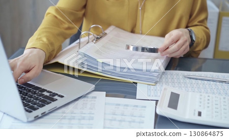 Female auditor in business attire inspecting financial reports with magnifying glass, surrounded by documents and a laptop and calculator. Business audit and taxes concept Female auditor in business attire inspecting financial reports with magnifying glass, surrounded by documents and a laptop and calculator. Business audit and taxes concept 130864875
