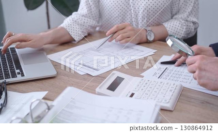 Female bookkeeper or auditor is working on finances papers with a colleague, using magnificent glass, calculator and laptop. Audit and taxes 130864912