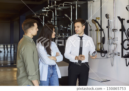 A salesman near the showcase of plumbing shop helping young couple to choose shower. 130865753