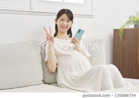 Pregnant woman with smartphone, peace sign, living room 130866754