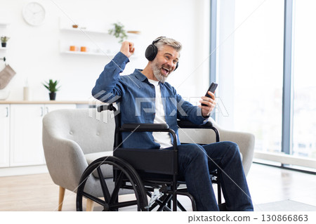 Mature Caucasian man, approximately mid-50s, sitting in wheelchair indoors, engaging with smartphone, wearing headphones, smiling enthusiastically, enjoying music, expressing happiness 130866863