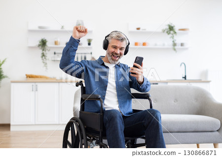 Mature Caucasian man in wheelchair using phone, listening to music with headphones, smiling and celebrating. Modern home setting with cozy interior. Concept of joy music and accessibility enhancement. 130866871