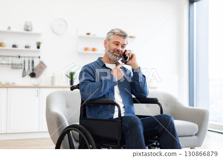 Mature Caucasian man with gray hair in wheelchair using phone, wearing denim and smiling in bright indoor setting, expressing happiness and independence 130866879