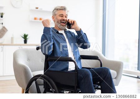 Mature Caucasian man sitting in wheelchair talking on smartphone, expressing excitement with clenched fists in modern living room environment 130866882