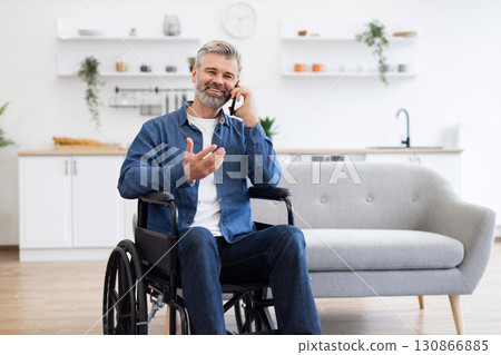 Mature Caucasian man with beard sitting in wheelchair in bright modern living room using mobile phone for communication 130866885