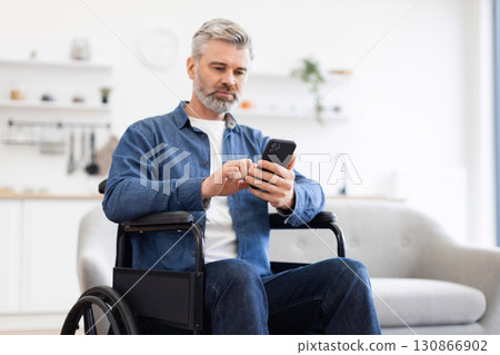 Mature Caucasian man seated in wheelchair using smartphone. Home setting with focus on accessibility, digital communication, and independence. Promotes themes of technology 130866902